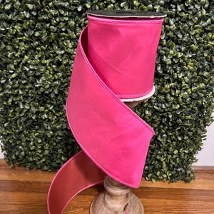 May include: A spool of solid hot pink ribbon with a smooth, matte finish. The ribbon is wrapped around a central core and cascades downward in a loose, flowing spiral, showcasing the vibrant, consistent color throughout the entire length.