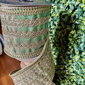 May include: A spool of wired ribbon featuring a metallic green base with a shimmering texture. The ribbon is decorated with three horizontal rows of intricate gold braided trim and finished with gold metallic edges along the top and bottom.