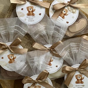 May include: A collection of small white organza bags tied with brown ribbon. Each bag contains a small white round tag with a brown teddy bear illustration and the text "Becky's Baby Shower 27/04/25".