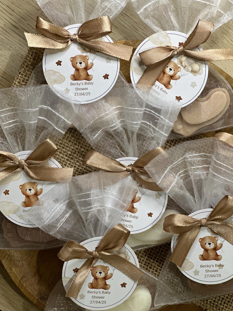 May include: A collection of small white organza bags tied with brown ribbon. Each bag contains a small white round tag with a brown teddy bear illustration and the text "Becky's Baby Shower 27/04/25".