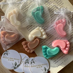 May include: A set of six baby foot-shaped soaps in pink, green, brown, and white, packaged in small organza bags. The soaps are arranged on a wooden surface with three round paper tags that say "Elouise's Baby Shower 27.07.24".