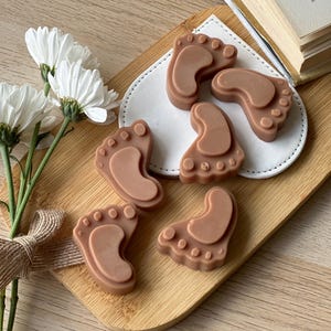 May include: Five brown baby foot-shaped soaps on a white plate, which is on a wooden surface. There are white flowers in the background.