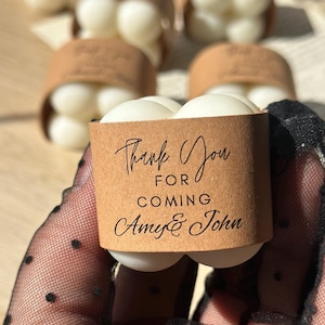 May include: A close-up of a gift box with white spheres and a brown paper wrap. The box has the text "Thank You For Coming Amy & John" in black script. The background shows more gift boxes.