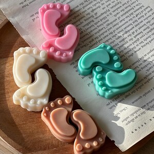 May include: Four pairs of baby foot-shaped soap bars in different colors: pink, white, light brown, and teal. The soaps are arranged on a wooden surface.