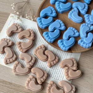 May include: Two sets of baby foot-shaped soaps, one set in light brown and the other in light blue. The soaps are arranged in a circular pattern on a wooden surface.