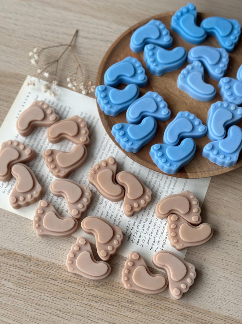 May include: Two sets of baby foot-shaped soaps, one set in light brown and the other in light blue. The soaps are arranged in a circular pattern on a wooden surface.