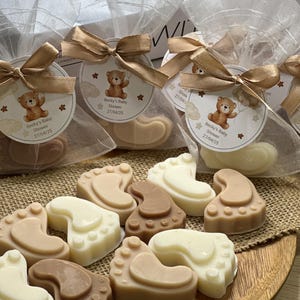 May include: Assortment of brown, beige, and white baby foot-shaped soaps wrapped in clear cellophane bags with brown ribbons and a small tag with a teddy bear and the text "Becky's Baby Shower 27/04/25".