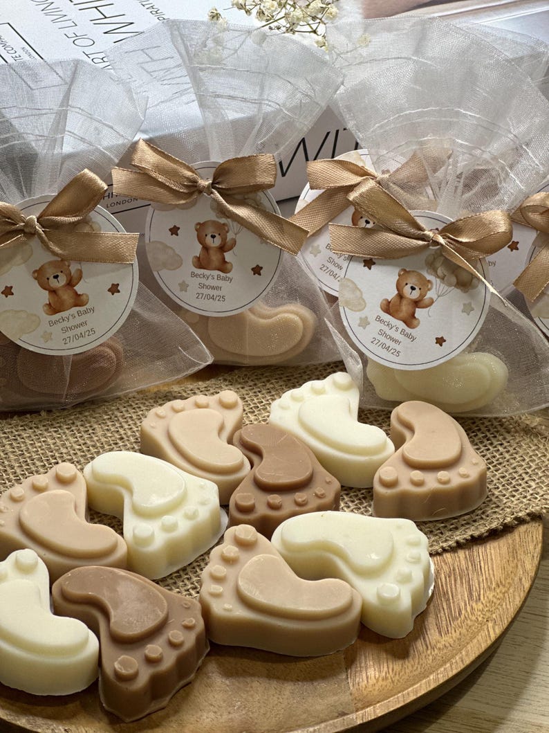 May include: Assortment of brown, beige, and white baby foot-shaped soaps wrapped in clear cellophane bags with brown ribbons and a small tag with a teddy bear and the text "Becky's Baby Shower 27/04/25".