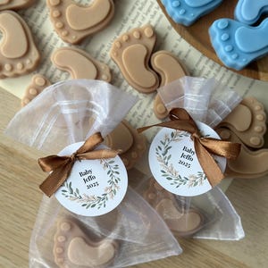 May include: Two white organza bags filled with small brown baby foot-shaped soaps and a round tag with the text "Baby Jeffo 2025" in a green wreath design.