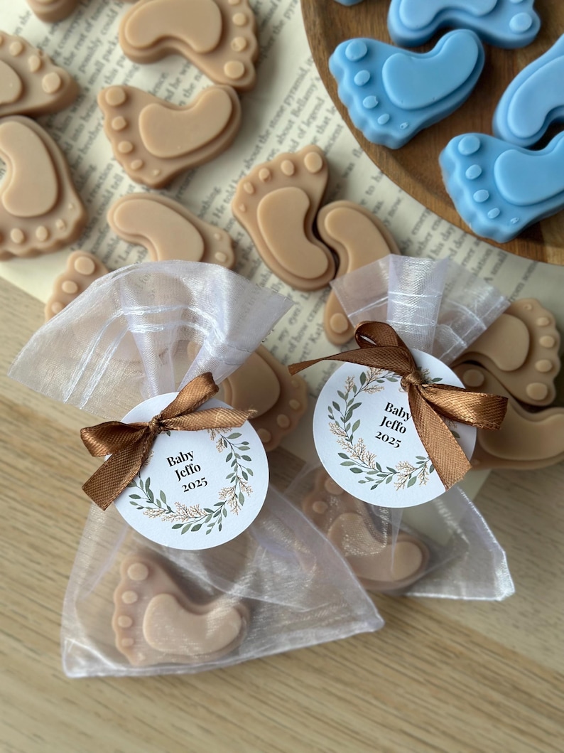 May include: Two white organza bags filled with small brown baby foot-shaped soaps and a round tag with the text "Baby Jeffo 2025" in a green wreath design.