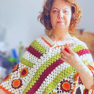 Pode incluir: Uma mulher vestindo um poncho de crochê com listras verdes, brancas, vermelhas e laranja. O poncho tem um design floral com flores de crochê.
