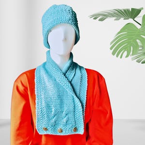 Pode incluir: Um chapéu e uma echarpe combinando em tricô azul turquesa com três botões dourados. O chapéu e a echarpe são usados por um manequim com uma camisa vermelha.