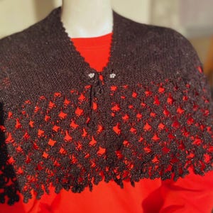 Pode incluir: Um xale de crochê preto com acabamento brilhante. O xale tem uma borda festonada e é amarrado no pescoço com um botão.