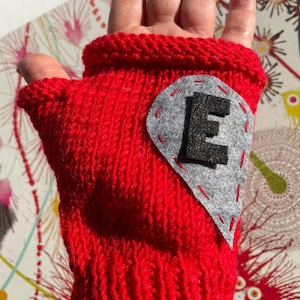 May include: Red knitted fingerless glove with a gray felt heart patch that has a black felt letter "E" on it.