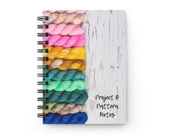 Pode incluir: Um caderno de espiral com um fundo de grão de madeira branca e uma pilha vertical de novelos de lã coloridos no lado esquerdo. O texto "Project & Pattern Notes" está impresso a preto no lado direito da capa.