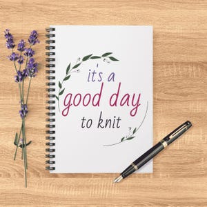 May include: A spiral-bound notebook with the text "it's a good day to knit" in purple and green. A black fountain pen and sprigs of purple lavender are on a light wood surface.