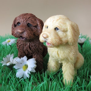 May include: Two small, realistic-looking dog figurines, one brown and one white, are sitting in a bed of green grass with white daisies.