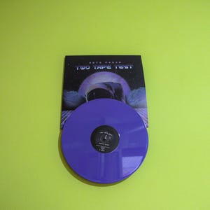 Peut inclure: Un disque vinyle violet avec le texte "TWO TAPE TEST" et le nom de l'artiste "ASTRO PARAS" sur la pochette. La pochette présente une image en noir et blanc d'un astronaute dans l'espace.
