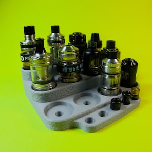 Puede incluir: Un organizador de plástico gris con ocho ranuras para guardar tanques de vape. El organizador tiene una superficie texturizada y está lleno de varios tanques de vape, incluidos tanques negros, plateados y transparentes.