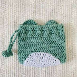 May include: A green crocheted drawstring bag with white crocheted ears and a white crocheted bottom. The bag has a drawstring closure.