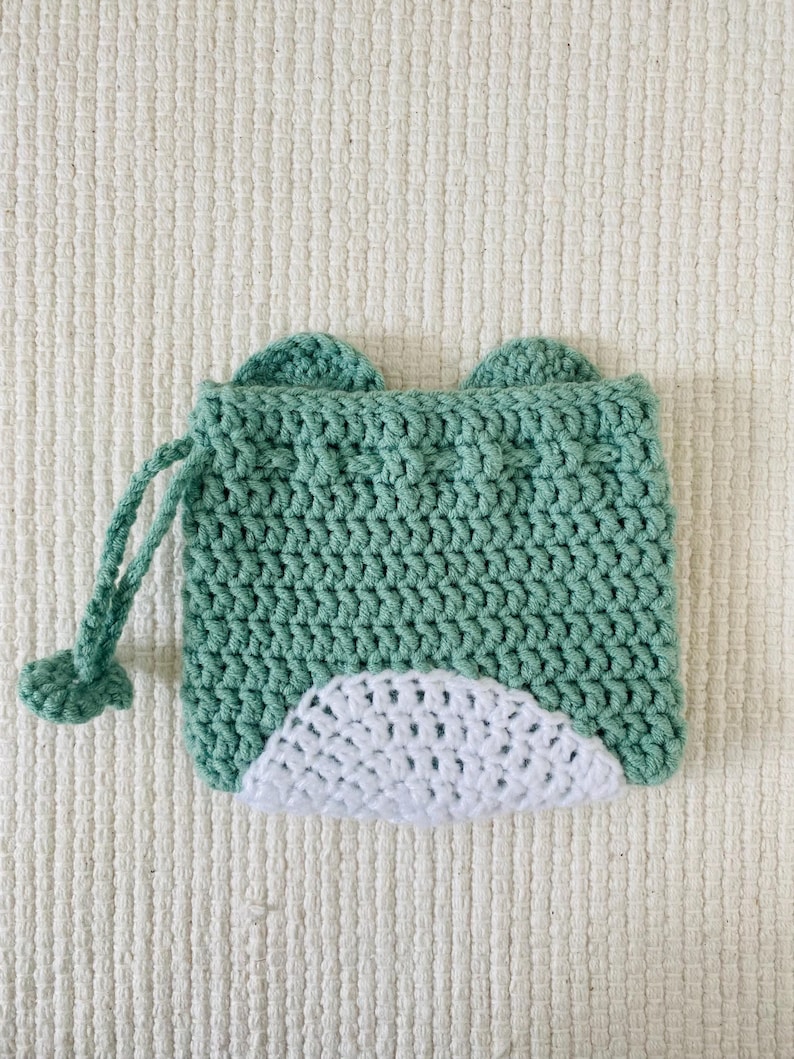 May include: A green crocheted drawstring bag with white crocheted ears and a white crocheted bottom. The bag has a drawstring closure.