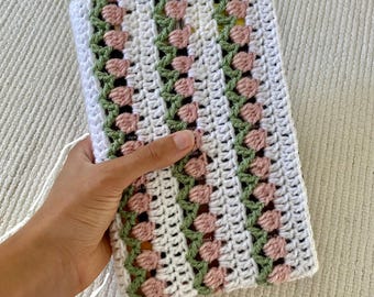 May include: A white crocheted book cover with pink and green floral accents. The cover is made with a textured stitch and has a simple, elegant design.