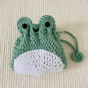 May include: A green crocheted frog-shaped drawstring bag with white crocheted accents and black button eyes.