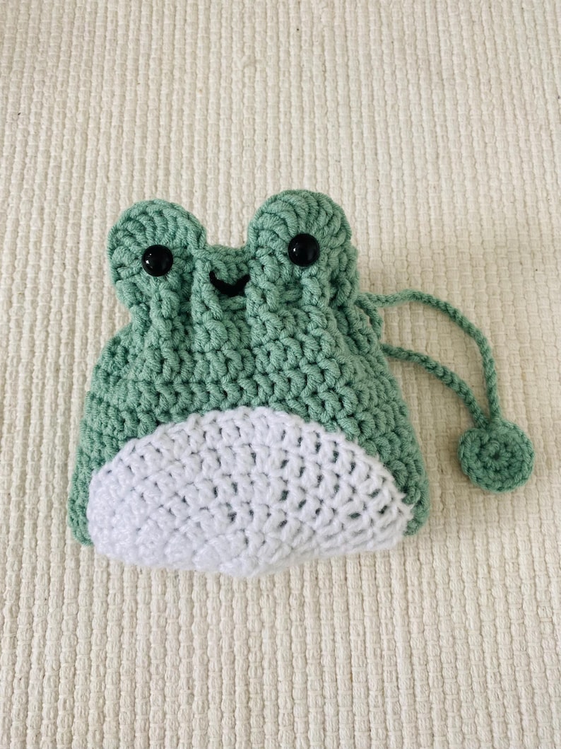 May include: A green crocheted frog-shaped drawstring bag with white crocheted accents and black button eyes.