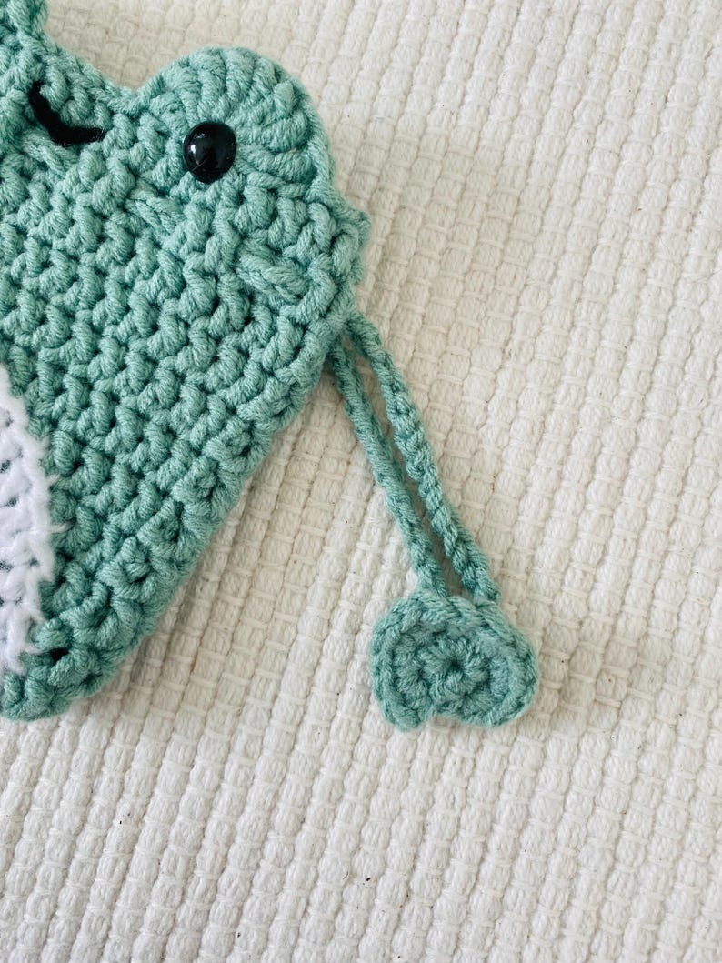 May include: A green crocheted pouch with a white crocheted detail and a black eye. The pouch has a green crocheted drawstring.