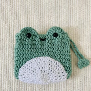 May include: A green crocheted frog-shaped pouch with a white crocheted belly and a drawstring closure. The pouch has black button eyes.