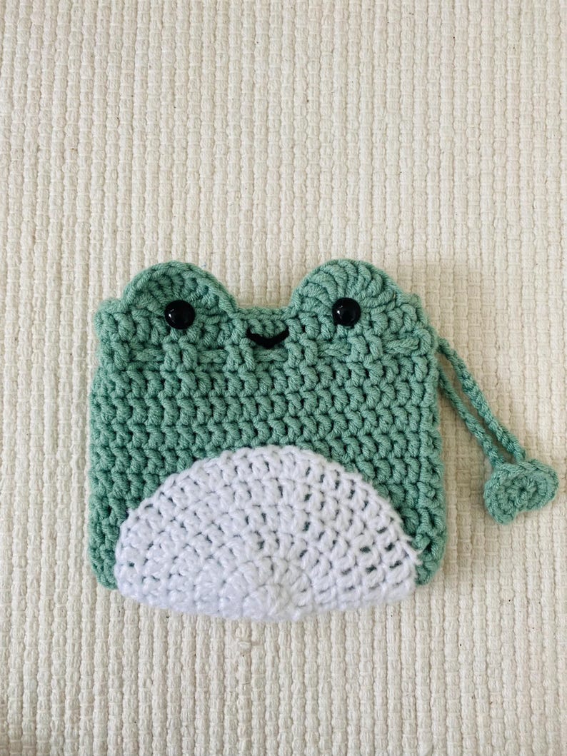 May include: A green crocheted frog-shaped pouch with a white crocheted belly and a drawstring closure. The pouch has black button eyes.