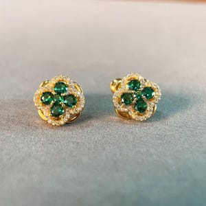 Könnte beinhalten: Ein Paar goldfarbene Ohrstecker. Jeder Ohrring hat ein vierblättriges Kleeblatt-Design mit grünen Edelsteinen in der Mitte und umgeben von kleineren, klaren Edelsteinen. Die Ohrringe sind vor einem hellgrauen Hintergrund platziert.