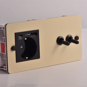 Könnte beinhalten: Eine beige Steckdose mit einer schwarzen Steckdose, USB-Anschlüssen und zwei schwarzen Schaltern. Die Steckdose ist auf einer weißen Kunststoffbasis mit rotem Akzent montiert.