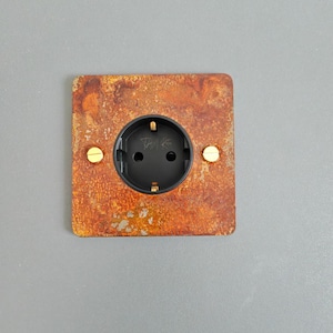 May include: A square-shaped electrical outlet with a black plastic cover and two gold screws. The outlet is mounted on a rusty metal plate.