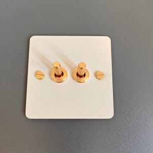 May include: A white light switch plate with two gold toggle switches. The switches have a textured, knurled design and are surrounded by a gold trim.
