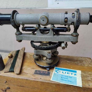 May include: A vintage silver and black surveying instrument with a wooden carrying case. The instrument has a telescope with a black lens and a graduated circle. The case has a label that reads "Lietz 820 Crown Road Burlingame, California 94010".