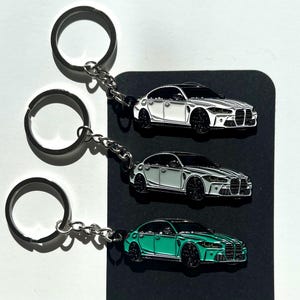 以下が含まれることがあります： エナメル加工された車のデザインが施された3つの金属製キーホルダー。2つは黒いホイールの白いセダン、1つは黒いホイールのティールカラーのセダンが描かれています。それぞれの車は、短いチェーンで銀色の金属製スプリットリングに取り付けられています。