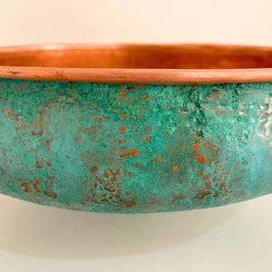 May include: A copper bowl with a green patina finish. The bowl has a rounded shape and a smooth, slightly textured surface.
