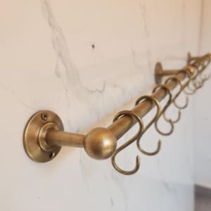 Könnte beinhalten: Eine messingfarbene Küchenreling mit mehreren Haken, montiert an einer weiß marmorierten Wand. Die Reling hat ein abgerundetes Design mit einer dekorativen Kugel am Ende. Die Haken sind zum Aufhängen von Küchenutensilien gedacht.