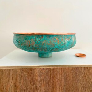 May include: A teal blue copper bowl with a copper rim and a small copper base. The bowl has a textured surface and is sitting on a wooden surface.