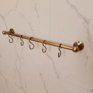 May include: A brass-colored, wall-mounted bar with six S-shaped hooks. The bar has decorative end pieces and is mounted on a white marble-patterned wall. This kitchen or bathroom accessory provides a stylish storage solution.