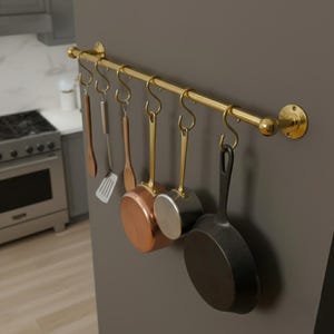 Peut inclure: Un rail de cuisine doré avec des ustensiles et des casseroles suspendus. Le rail est fixé à un mur gris et comprend une cuillère en bois, une spatule, des casseroles en cuivre et une poêle en fonte noire.