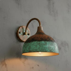 Może przedstawiać: Rustykalna lampa ścienna z miedzianym wykończeniem. Klosz w kształcie kopuły ma zieloną i brązową patynę oraz zakrzywione ramię. Światło jest zamontowane na okrągłej płycie tylnej. Wnętrze klosza jest złote.