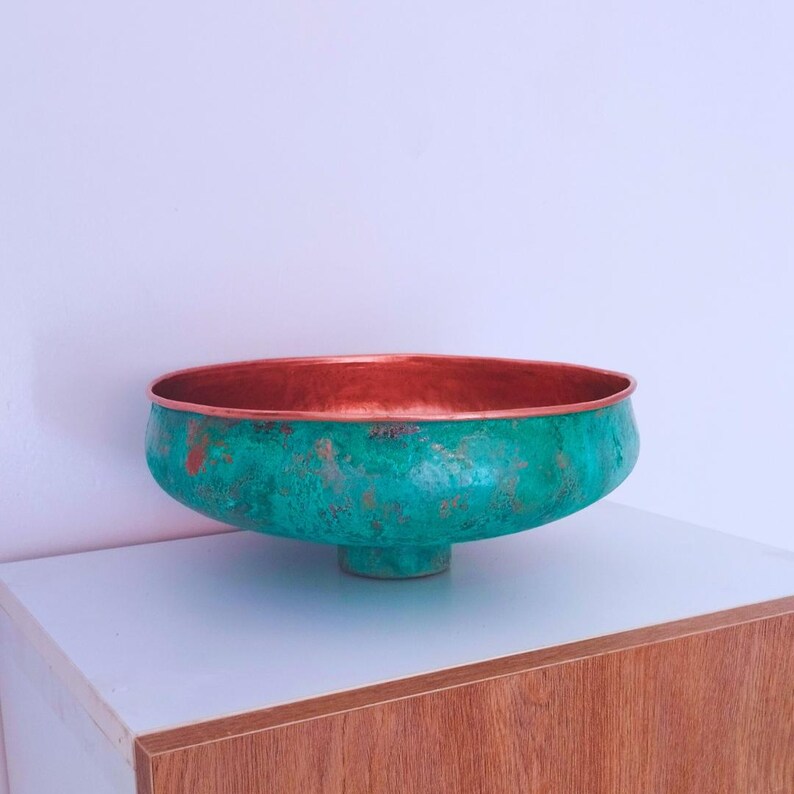 May include: A large, copper bowl with a green patina finish. The bowl has a wide, round base and a flared rim. The bowl is sitting on a wooden surface.