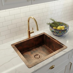 May include: A copper kitchen sink with a hammered texture, featuring a gold faucet. A decorative bowl with lemons and herbs sits nearby. The sink is set into a white countertop with white cabinets and a white tile backsplash.
