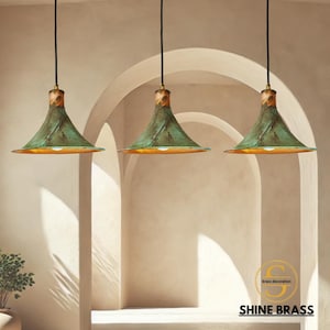 Puede incluir: Tres lámparas colgantes con una pantalla verde texturizada y un interior dorado. Las luces cuelgan de cables negros y tienen un acento de madera en la parte superior. El texto "SHINE BRASS" es visible en la parte inferior.