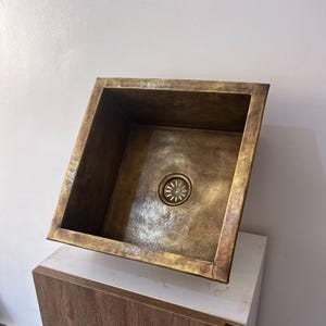 May include: A square, antique-style copper sink with a bronze finish. The sink has a central drain and a slightly textured surface. The edges are angled, and the overall design is rustic and elegant.