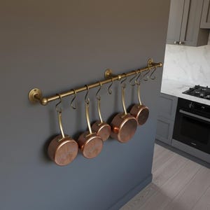Peut inclure: Un porte-casseroles en laiton avec six casseroles en cuivre suspendues. Le support est fixé à un mur gris. Les casseroles ont des fonds en cuivre et des poignées en laiton. La cuisine comprend des placards et une cuisinière.