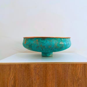 May include: A teal green copper bowl with a copper rim and a pedestal base. The bowl has a distressed, aged look.