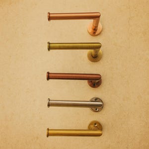 May include: Five metal toilet roll holders in copper, brass, and silver finishes. Each has a horizontal bar and a circular base for wall mounting. The holders are arranged vertically against a light-coloured wall.